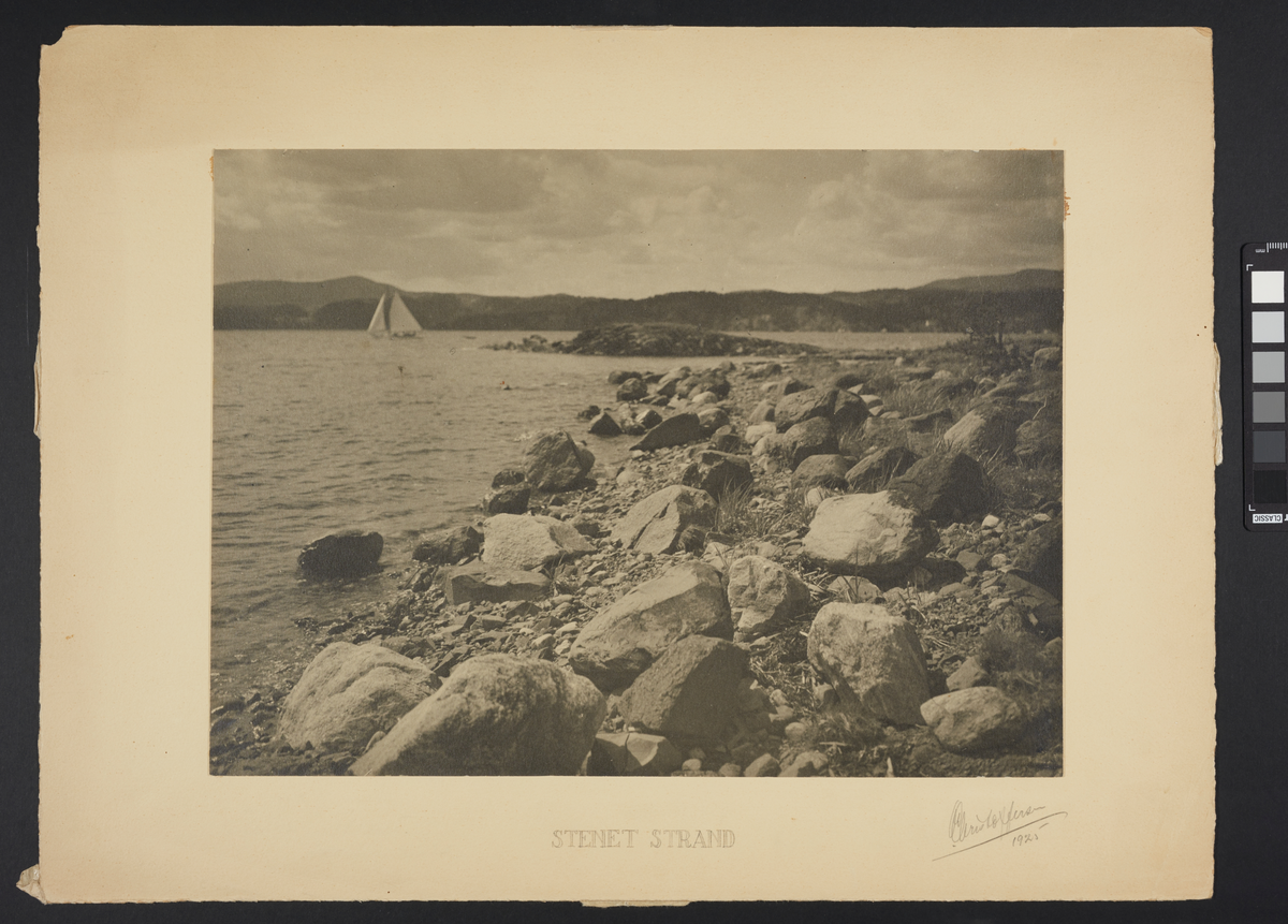 En strand ved sjøen som er dekket av store steiner. Ute på vannet ved horisonten er det en seilbåt, og bak den strekker det seg en landmasse med myke fjell.