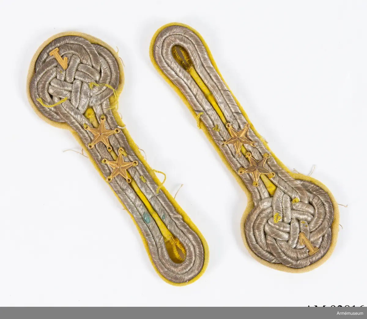 Ett par axeltränsar i silver på gul botten med guldfärgade metalldekorationer.

Axeltränsarna är gjorda av dubbelt silverfärgat beläggningssnöre av metalltråd i en platt flätad form med tre dubbla knutor och en stjälk vars ögla bildar ett knapphål. Ovanpå den flätade formen är fastsytt två femuddiga stjärnor samt en 1:a i mässing.
Allt samman är sedan fastsytt på en botten av gult kläde i vars kanter syns rester av oblekt lingarn som visar att tränsarna sprättats loss från axlarna på en uniform.