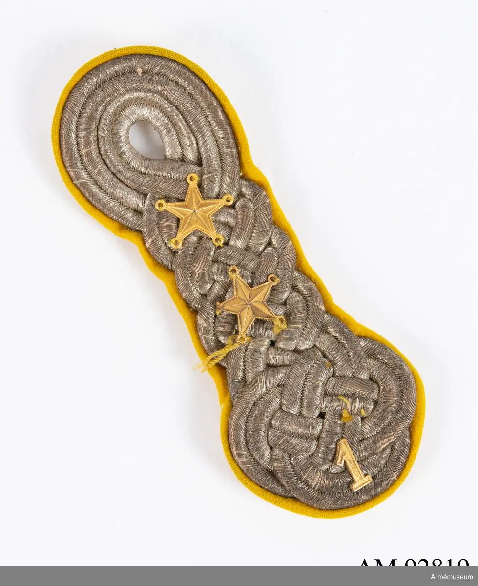 Axelträns i silver med guldfärgade dekorationer på gul botten.

Axeltränsen är gjord av dubbelt silverfärgat beläggningssnöre av metalltråd i en platt flätad form med en tredubbel knut nederst, ovanför detta tre dubbla flätningar och en ögla som bildar ett knapphål. Ovanpå denna flätade form är fastsytt två femuddiga stjärnor samt nederst en 1:a och kung Gustaf V:s (förgyllda?) namnchiffer i mässing.
Allt samman är sedan fastsytt på en botten av gult kläde.