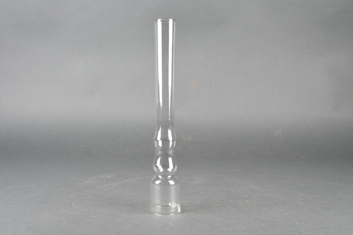Oljelampe bestående av stativ av metall, skjerm av blikk malt grønn på oversiden og hvit på undersiden, parafinbrenner av glass, metall og veke av vevet tekstil, samt et lampeglass.