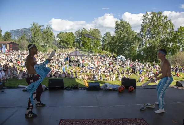 To unge menn står på en scene foran et stort publikum. De sjonglerer kjegler mellom seg