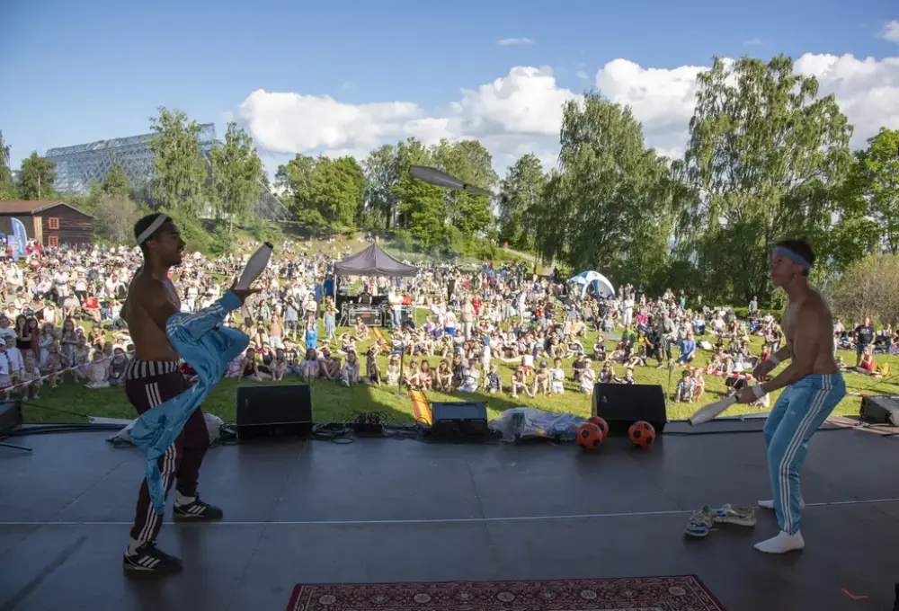 To unge menn står på en scene foran et stort publikum. De sjonglerer kjegler mellom seg