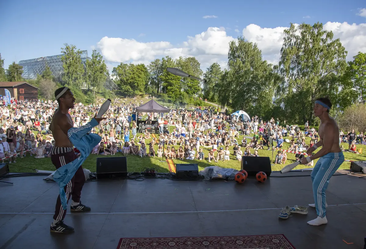 To unge menn står på en scene foran et stort publikum. De sjonglerer kjegler mellom seg