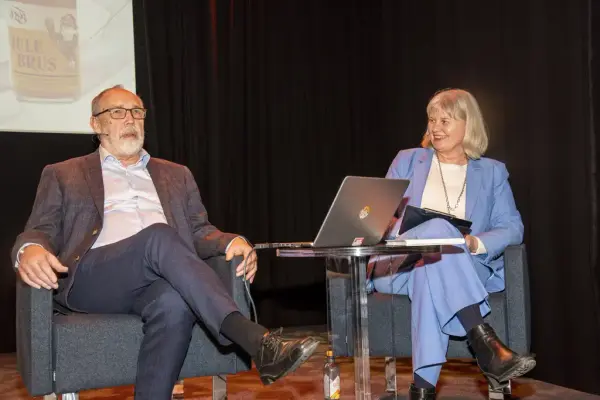 En eldre mann og en kvinne sitter i hver sin stol foran en storskjerm og snakker. De har et bord med en laptop i mellom seg.