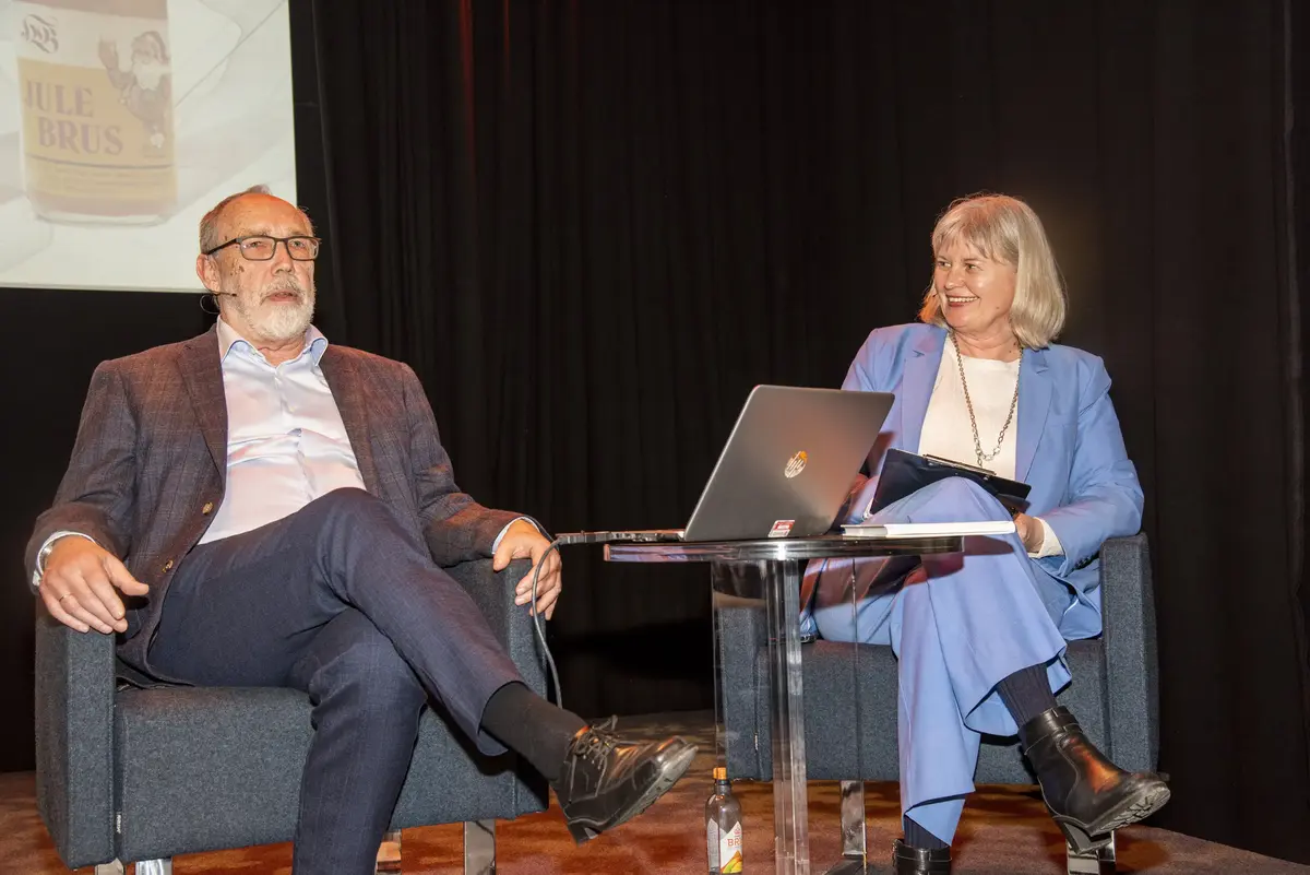 En eldre mann og en kvinne sitter i hver sin stol foran en storskjerm og snakker. De har et bord med en laptop i mellom seg.