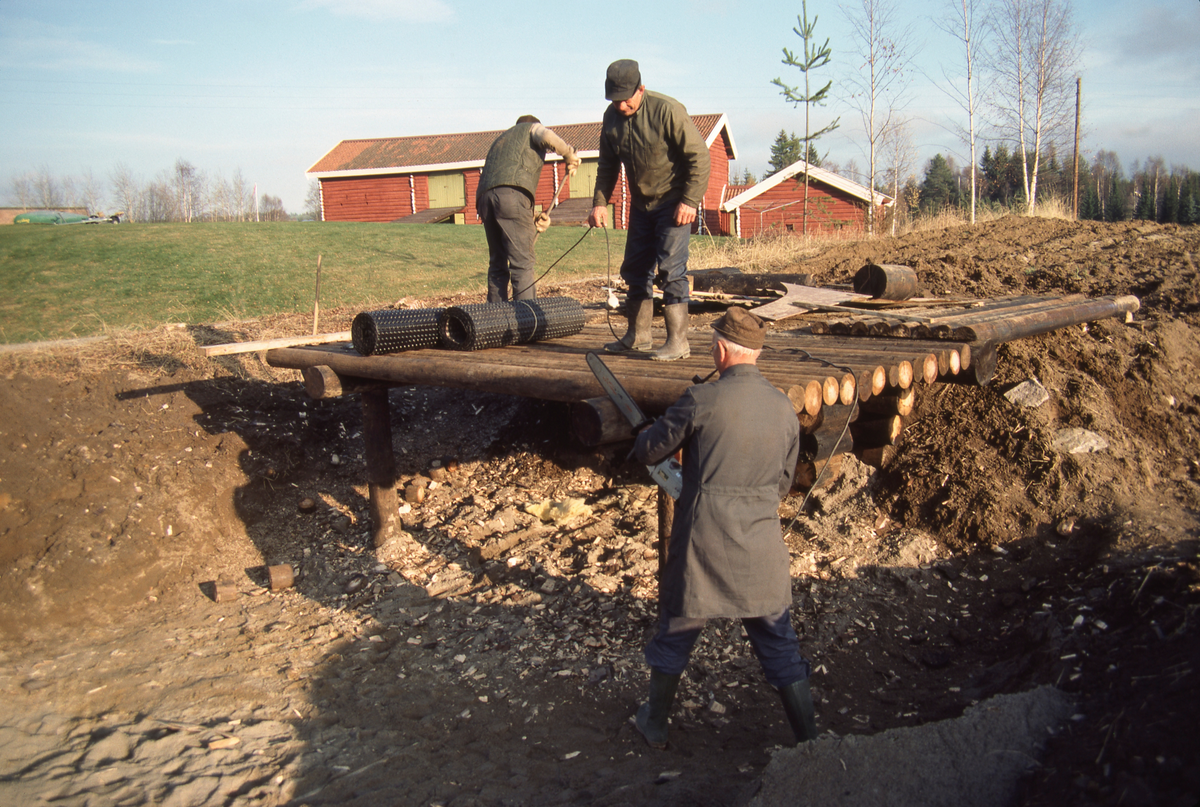 Fra bygginga av en blesterovn – etter modell fra Ole Olsen Evenstads hefte «Afhandling om Jern-Malm som findes i Myrer og Moradser i Norge, og Omgangsmaaden med at forvandle den til Jern og Staal» (1782/1790) – på Norsk Skogbruksmuseum høsten 1984. Fotografiet er tatt mot det som skulle bli fronten på ovnen, der det var bygd et golv av kreosotimpregnert tømmer, båret av to stolper. På golvet lå det to ruller med grunnmursplast, som antakelig skulle legges på det nevnte golvet, som deretter skulle dekkes med jord. Under golvet skimter vi et av hjørnene i den laftete treramma som omgav det Evenstad kalte «Blæsteren», sjølve ildstedet. De to store blåsebelgene som skulle pumpe oksygenrik luft inn i ildstedet skulle plasseres under plattformen i forgrunnen, med munnstykker av jern i ei trang sjakt i botnen av ovnen. Da dette fotografiet ble tatt sto tre av medarbeiderne i museets driftsavdeling på golvet. I forgrunnen, med ryggen mot fotografen, sto forvalter Håkon Sæle, kledd i lagerfrakk, med pipa i munnen og med ei motorsag i hendene. Han kappet enden på ei av lunnene som bar golvet. Oppå plattformen ser vi to menn – den av dem som sto vendt mot fotografen var antakelig hjelpearbeider Jakob Nersveen. Bygningene i bakgrunnen er stallåven og sommerfjøset på klokkergarden Fossum.

I Norsk Skogbruksmuseums årsmelding for 1984 står dette om bygginga av blesterovn på museet: «En jernblesterovn for brenning av jern av myrmalm ble bygget i oktober/november. Blesterovnen er bygget etter Ole Olsen Evenstads tegninger i hans bok om myrmalmbrenning, utgitt i 1790. Lærer Ivar Berre fra Skogn har erfaring med jernutvinning i blesterovn, og har vært både konsulent og deltatt i oppstarting av arbeidet. Prosjektet er ellers gjennomført av museets egne håndverkere.»