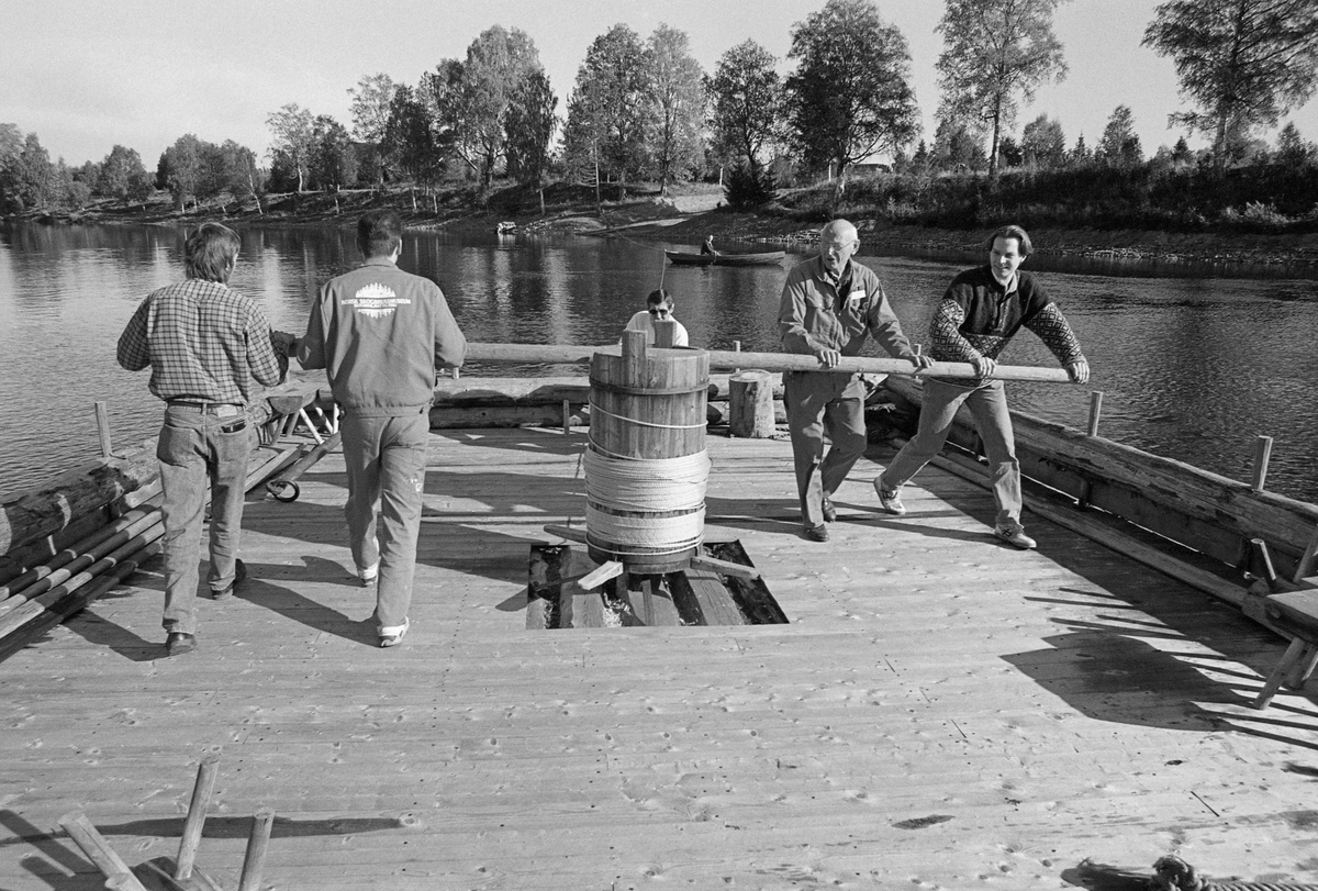 Norsk Skogbruksmuseums rekonstruerte spillflåte, som fra sommeren 1993 og flere år framover lå forankret i lensesteinen i Glåma, like ovenfor Prestfossen og Klokkerfossen, «spilles» i land for vinterlagring. Dette arbeidet ble gjort av museets personale. Karene som skjøv armene på gangspillet var Kjell Arne Grøtbekklien og Kjell Magne Kaveldiget fra driftsavdelingen (til venstre, med ansiktene mot fotografen) og Håkon Sæle og Bjarte Wigdel (til høyre, med ansiktet mot fotografen). Fotografiet ble tatt høsten 1994.