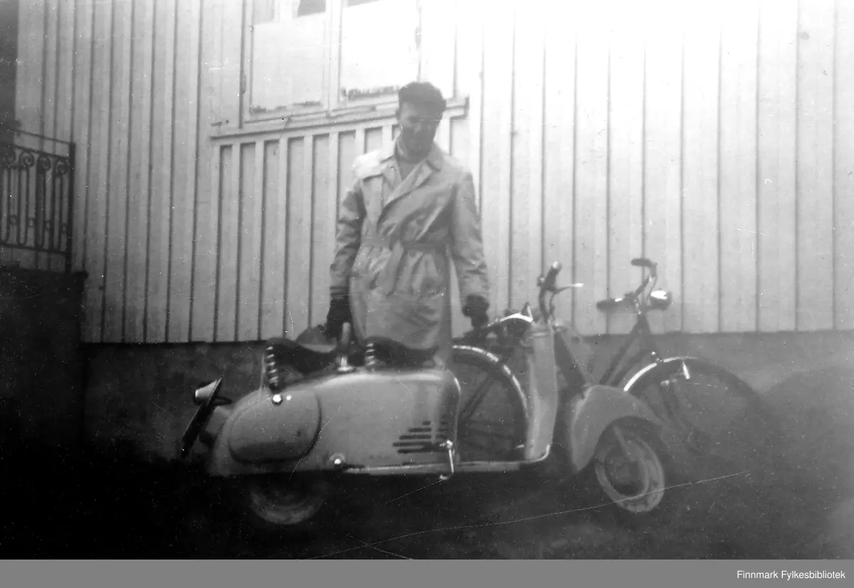 Ingar Bjørnstad med ny Puck scooter utenfor doktorboligen i Havøysund, 1955. En sykkel lener mot veggen.