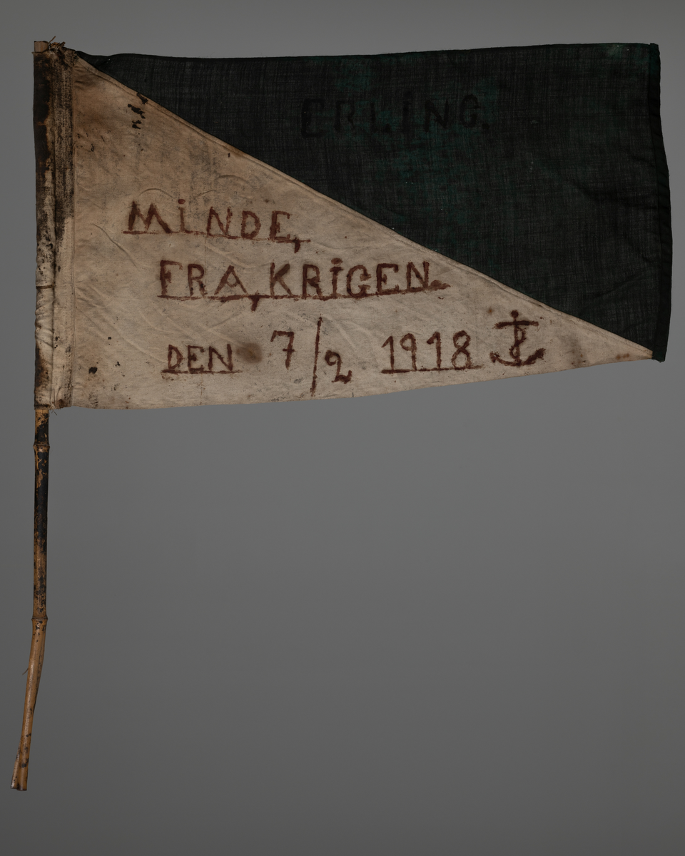 Flagg i lin festet til bambusstang. Bambusstangen er sprekt/ flisete i enden. Flagget er sydd rundt stanga. Flagget er todelt, delt diagonalt. Øverste del er malt grønn. Navnet "Erling" er malt på. Nederste del er ufarget (naturhvit). Tekst malt på: "Minde fra krigen den 7/2 1918".
