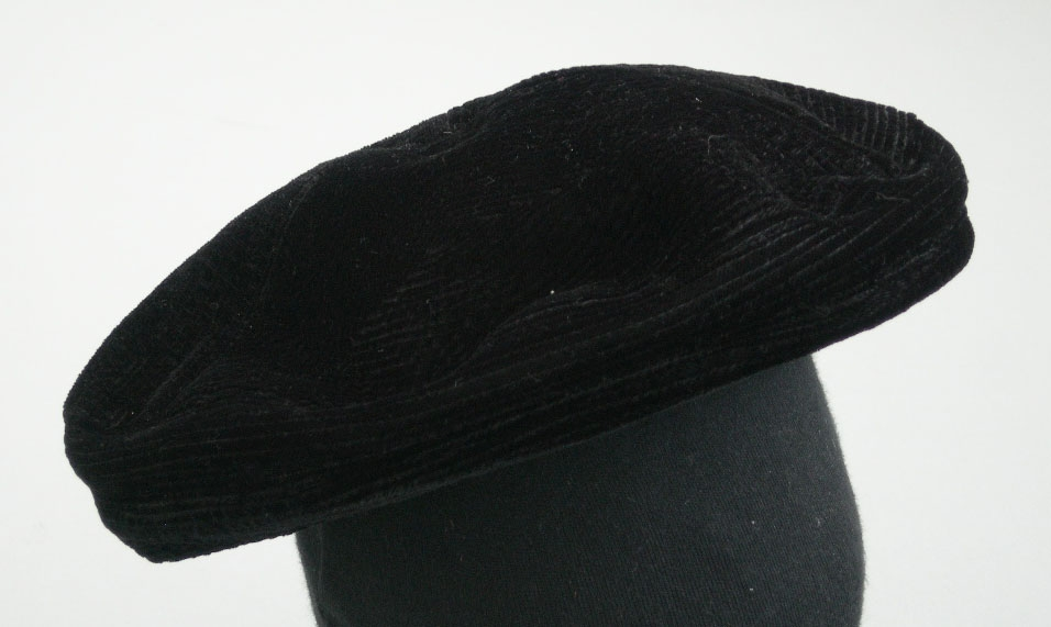Svart basker av konstsilke och sammet med platt kulle sydd av tre stycken. Kulle och kant med täta stickningar. Taftfoder, ripsband innanför kanten.
