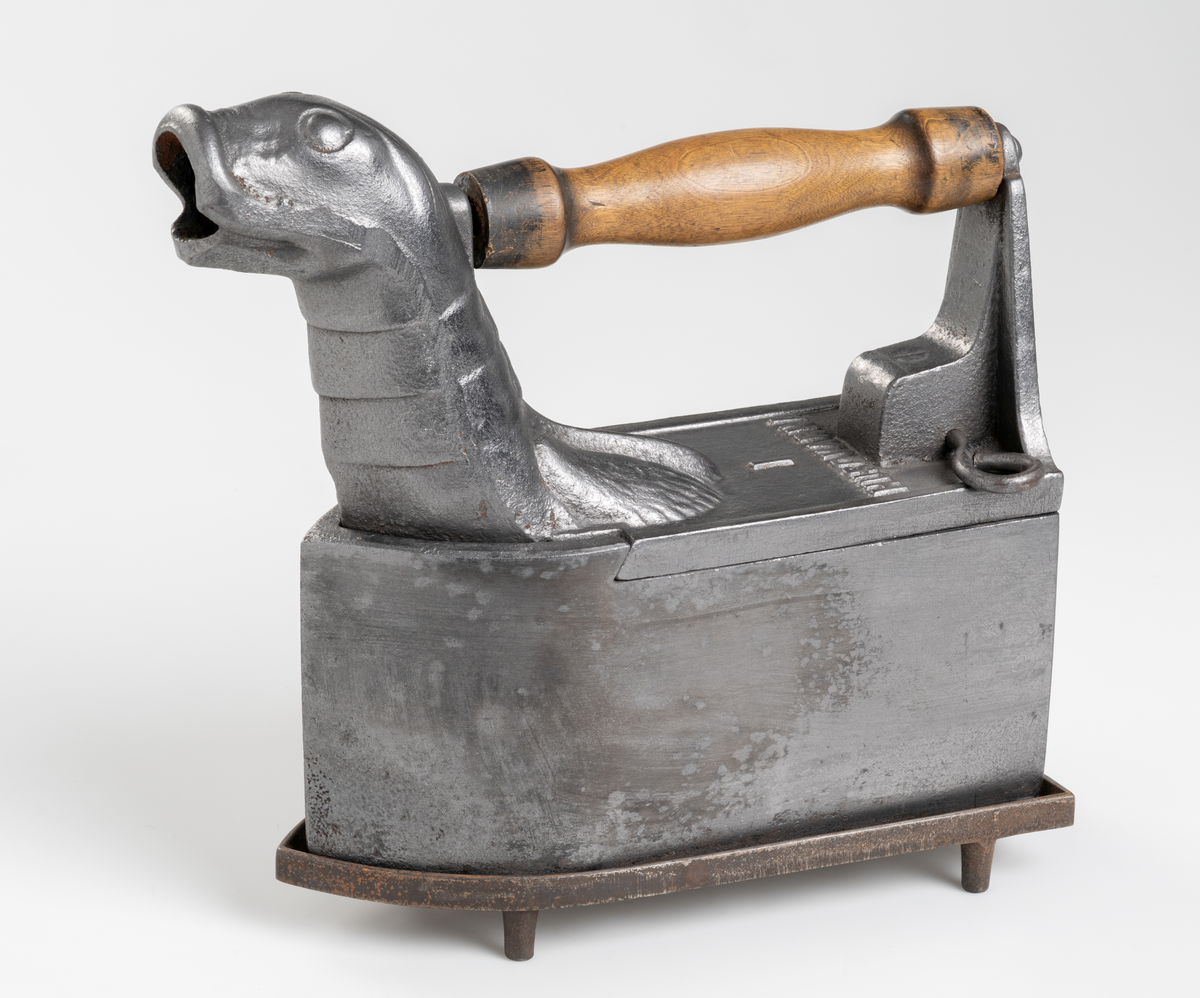 Dekorativt strykjärn, pressjärn, av järn med kolbehållare. Utformat med djurhuvud. Handtag av trä. Tillhörande strykjärnsställ av järn.