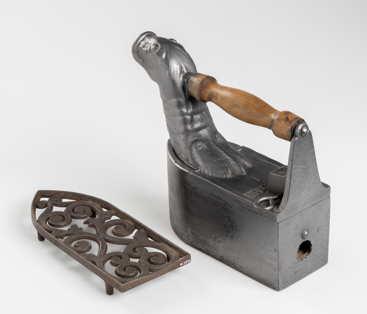 Dekorativt strykjärn, pressjärn, av järn med kolbehållare. Utformat med djurhuvud. Handtag av trä. Tillhörande strykjärnsställ av järn.