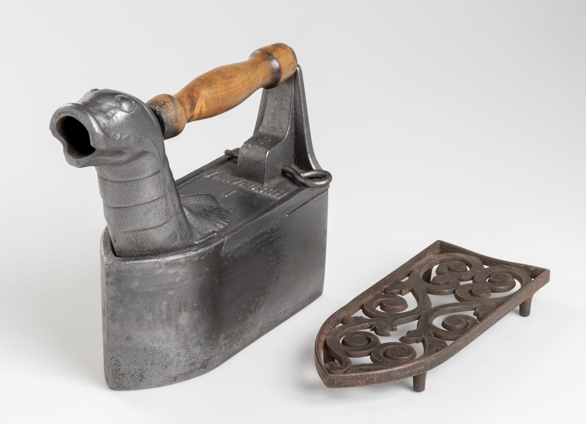 Dekorativt strykjärn, pressjärn, av järn med kolbehållare. Utformat med djurhuvud. Handtag av trä. Tillhörande strykjärnsställ av järn.
