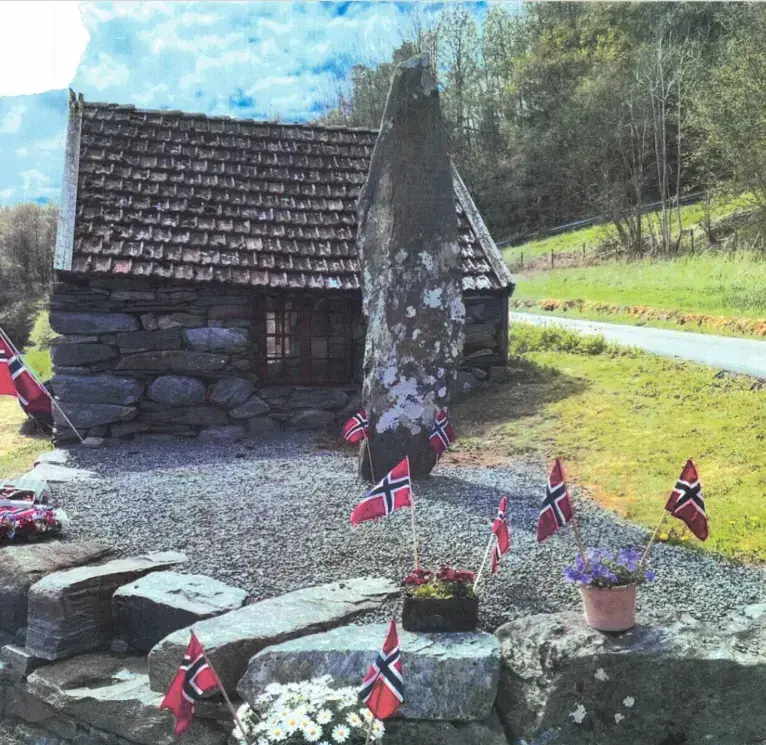 Bautastein foran et lite hus bygget i stein. Steinmur med blomsterpotter på, pyntet med norske flagg.