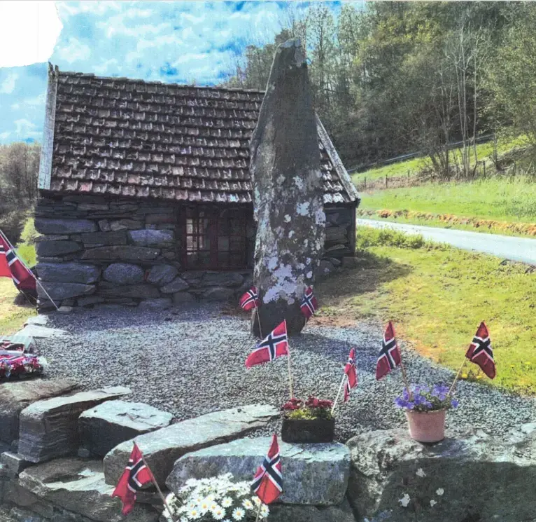 Bautastein foran et lite hus bygget i stein. Steinmur med blomsterpotter på, pyntet med norske flagg.