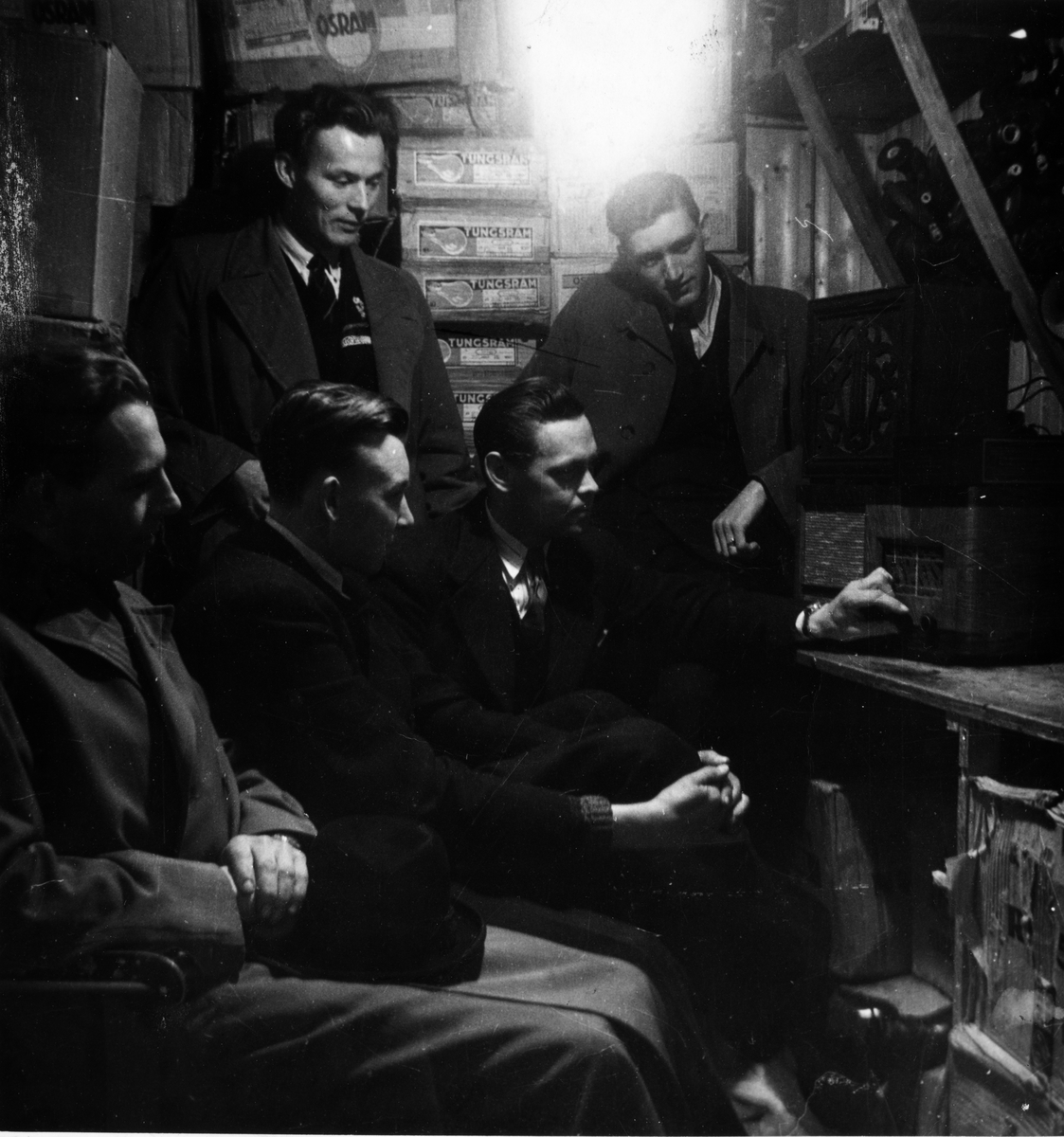 Tyske "Wehrmacht" har kapitulert - dagen er 8. mai 1945. Bildet er fra radiolytterommet hos O. Hollingsæter el. teknisk forretning i Kristiansund. Her fikk en stemmen fra London under hele krigen. Rommet var lydisolert med lampekartonger - bl.a. fra Tungsram. Fra venstre: Rolf Moen (elektriker), Tor Thoresen (læregutt), Kåre Hagedal (lagersjef), el. tekniker Steene og snekker Torbjørn Gjeldnes. Apparatet vi ser er en Tandberg batteriradio. Fra en samling gitt til museet 08.04.2025 av Eivind Thoresen.