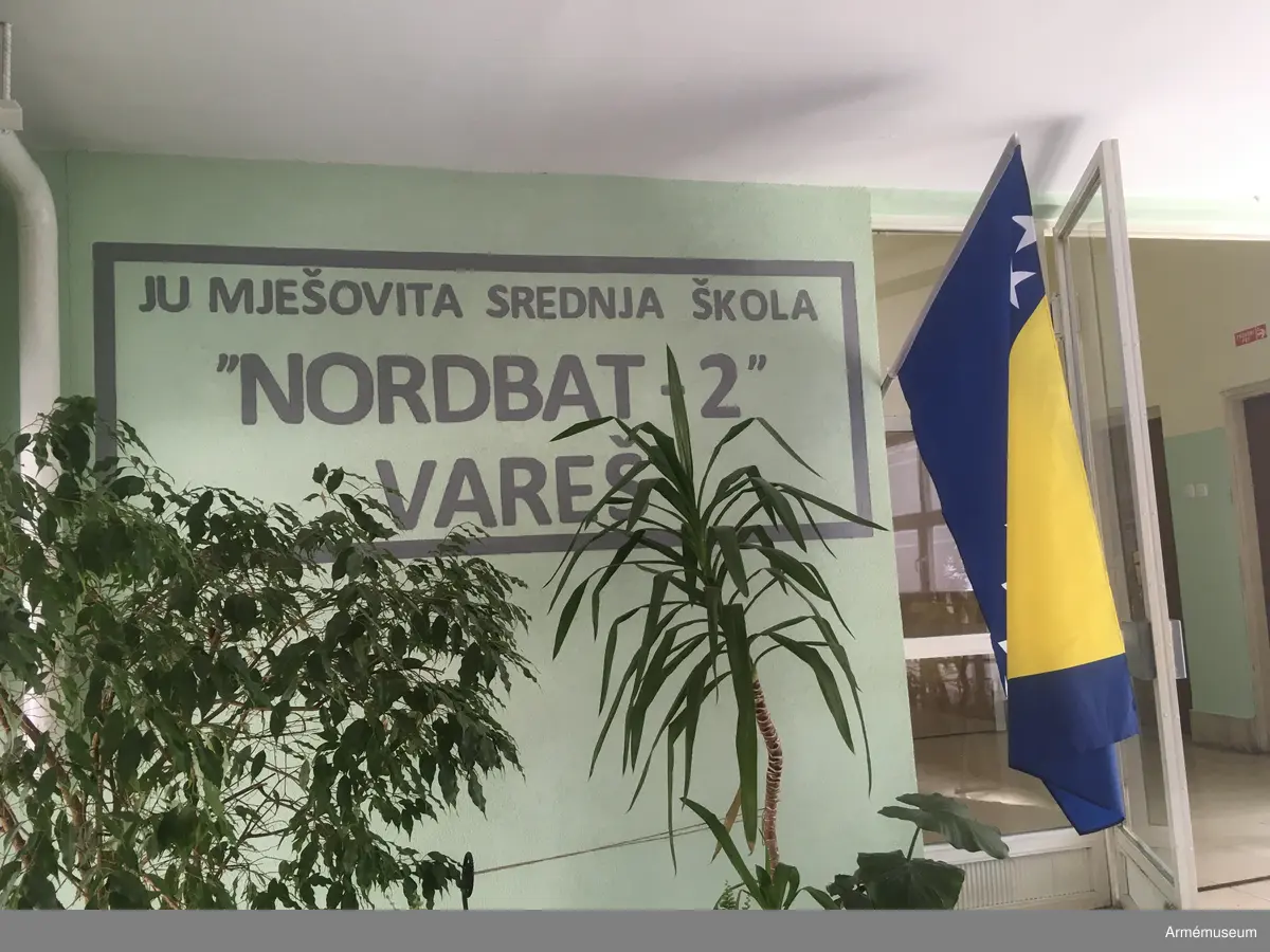 Metallplatta med inskription som varit fäst på insidan av ett fönster på gymnasieskolan Goran Kovacic i Vares i Bosnien. Under 1994 döptes skolan om till ”Nordbat 2” efter de händelser som tidigare tilldrog sig i och omkring skolan där delar av den svensk-danska pansarskyttebataljonen som ingick i UNPROFOR befann sig. Nordbat 2 ingick i BA01 som var den första av de svenska bosnienbataljonerna för de fredsbevarande styrkorna. 

På baksidan av plattan återfinns inskriptioner från elever på skolan. Föremålet förmedlades genom överste Ulf Henricsson och Stig-Inge Blennow.