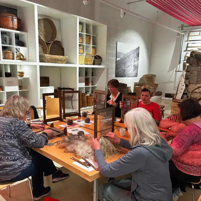 Folk på kurs i veving av minivarafell sitter rundt eit bord og vever