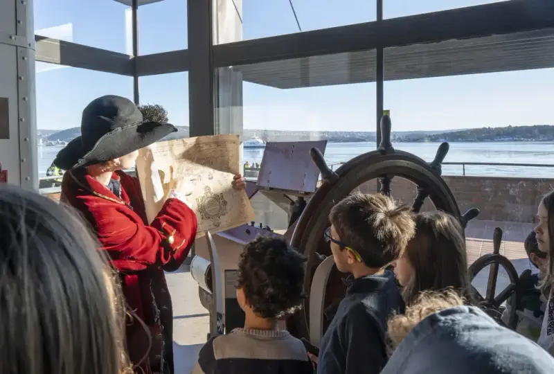 Person utkledd som pirat viser et skattekart til en gruppe barn i museet, med Oslofjorden i bakgrunnen.