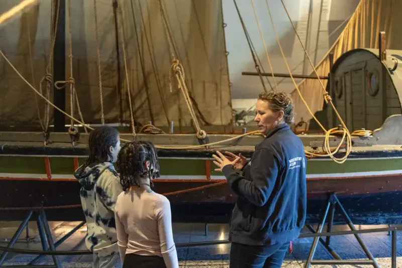 To barn og en formidler foran en tradisjonsbåt i utstillingen «Sjøveien» på Norsk Maritimt Museum.