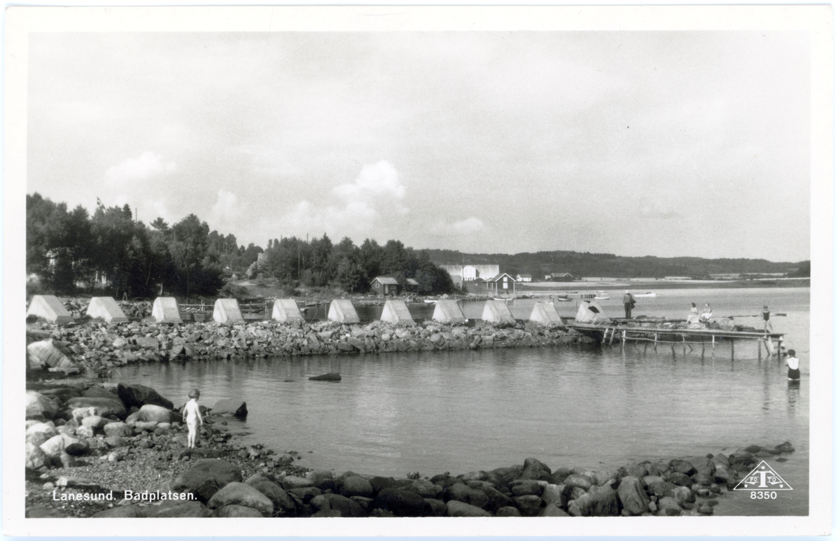 Vykort som avbildar landskap vid havet. Människor badar till höger i bild vid brygga, ett barn står i strandkanten.