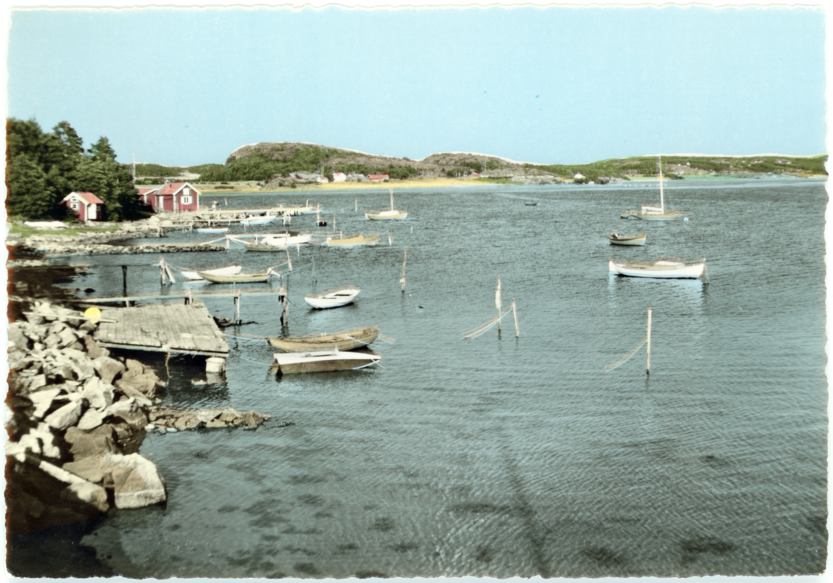 Handkoloreratvykort som avbildar landskap vid havet; bryggor sträcker sig ut i havet där småbåtar ligger, med berg i motivets bakgrund.