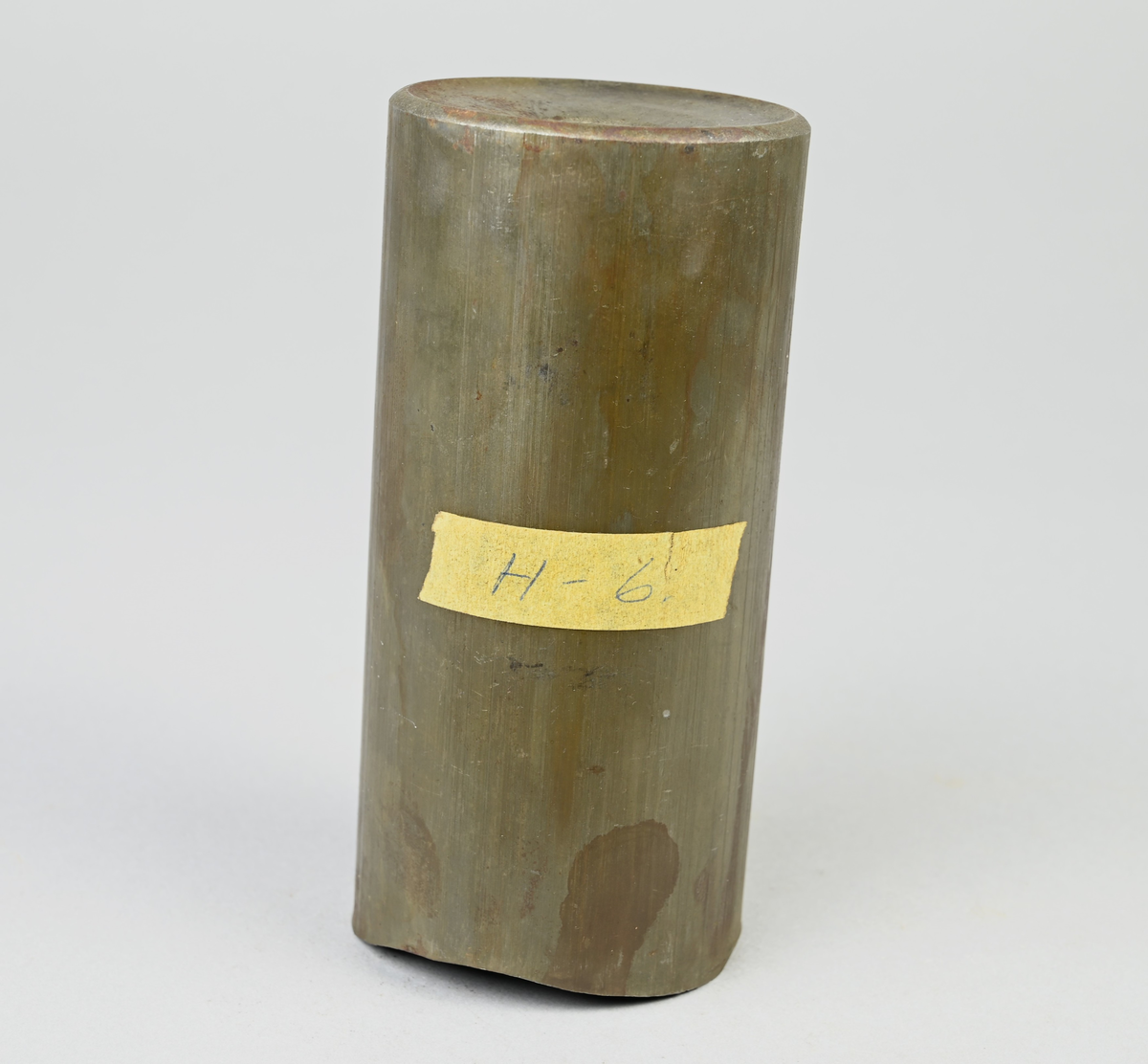 Prov på projektilhylsa. Del av tillverkningsserie. Pressad av stål från runt plåtämne vid Skultuna bruk. Märkning på provet med tejbit med notering: "H-6", samt med etikett med notering: "716".