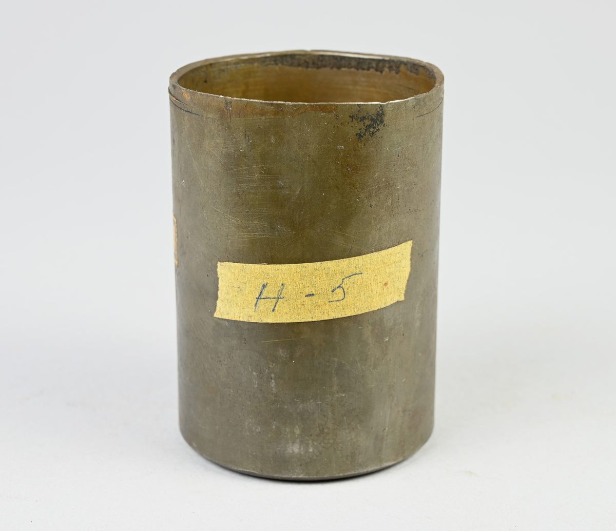 Prov på projektilhylsa. Del av tillverkningsserie. Pressad av stål från runt plåtämne vid Skultuna bruk. Märkning på provet med tejbit med notering: "H-5", samt med etikett med notering: "716".