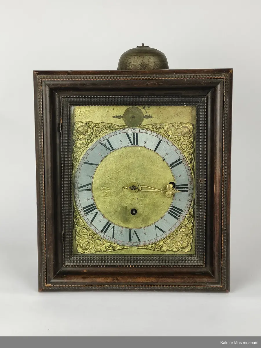 KLM 20535. Väggur. Fyrkantigt fodral av brunbetsad björk dekorerat med hopplister med lucka på sidorna och glasdörr framför urtavlan. Förgylld urtavla med hörnornament i förgyllt bly. Sifferring i tenn. Upptill en rund liten tavla i mässing med signatur: Johan Kock Holmiae 1672. Även urverket är signerat. Urverk av mässing med slagverk. Har haft lod och trissor som numera saknas.