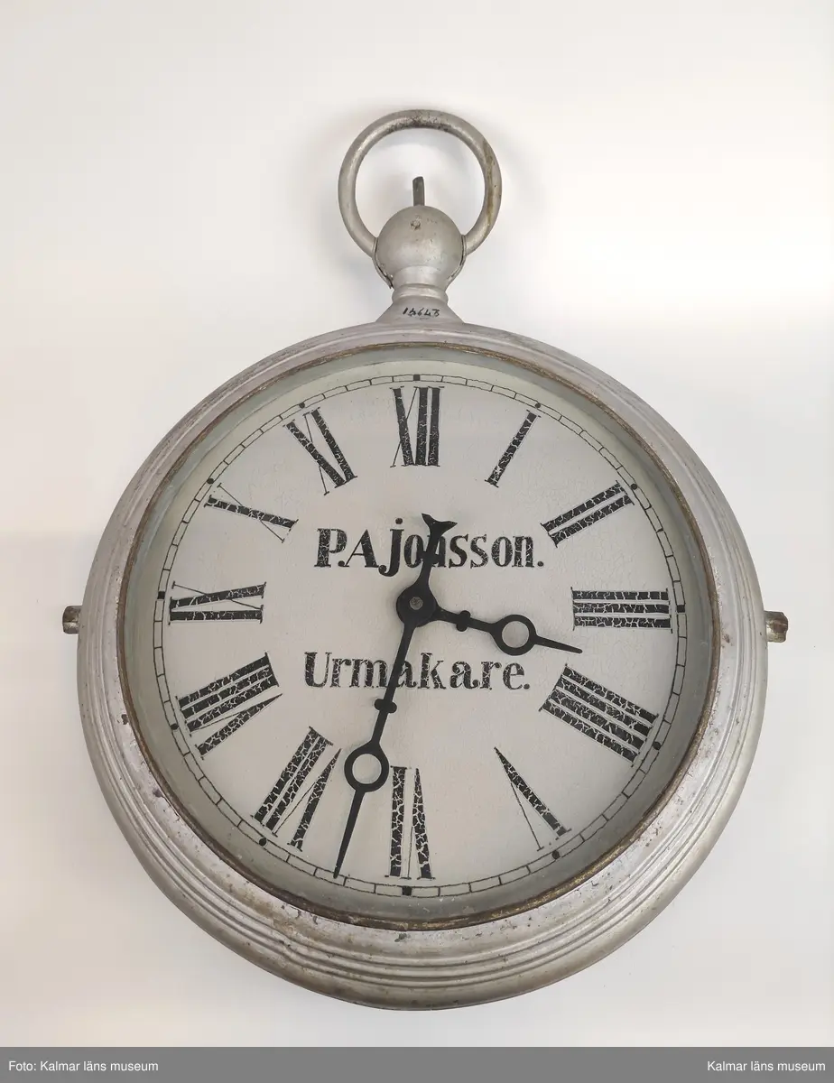 KLM 27941. Skylt. Av metall målad i silverfärg samt glas. Urmakareskylt i form av ett stort fickur. Målad urtavla med vit bakgrund och svarta romerska siffror samt text: P.A. Jonsson urmakare.