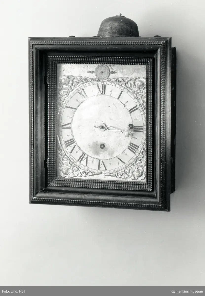 KLM 20535. Väggur. Fyrkantigt fodral av brunbetsad björk dekorerat med hopplister med lucka på sidorna och glasdörr framför urtavlan. Förgylld urtavla med hörnornament i förgyllt bly. Sifferring i tenn. Upptill en rund liten tavla i mässing med signatur: Johan Kock Holmiae 1672. Även urverket är signerat. Urverk av mässing med slagverk. Har haft lod och trissor som numera saknas.