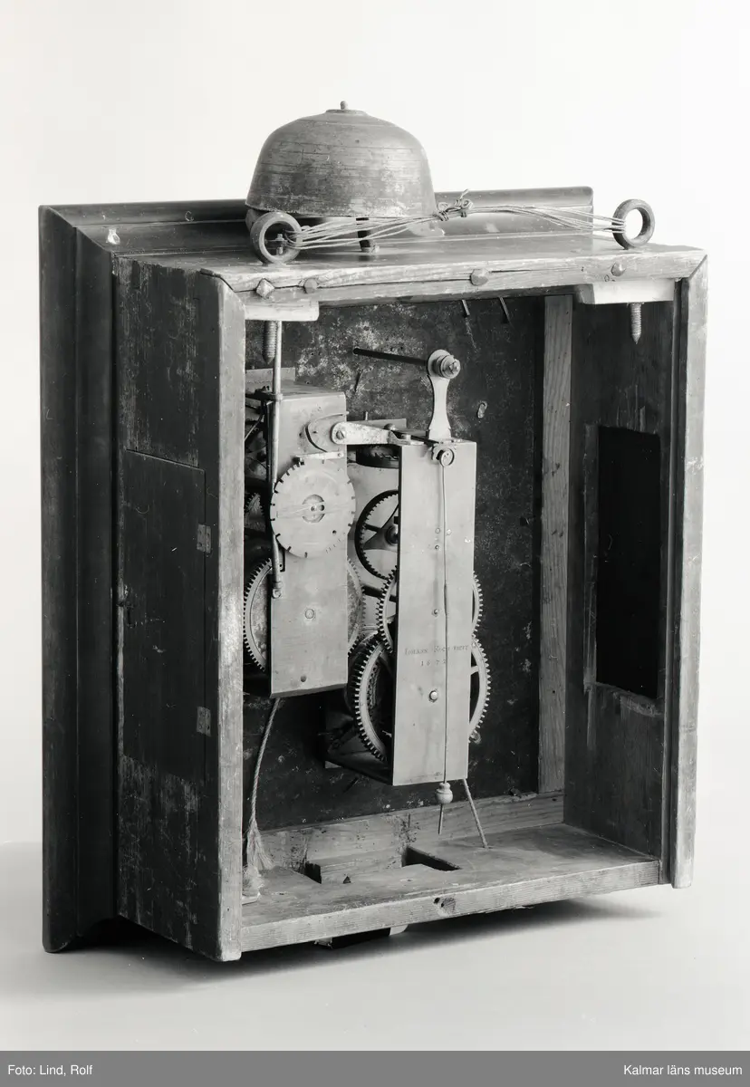 KLM 20535. Väggur. Fyrkantigt fodral av brunbetsad björk dekorerat med hopplister med lucka på sidorna och glasdörr framför urtavlan. Förgylld urtavla med hörnornament i förgyllt bly. Sifferring i tenn. Upptill en rund liten tavla i mässing med signatur: Johan Kock Holmiae 1672. Även urverket är signerat. Urverk av mässing med slagverk. Har haft lod och trissor som numera saknas.