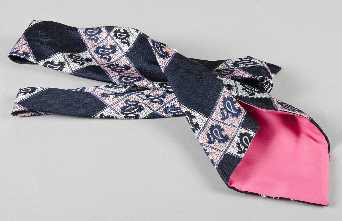 Slips av syntetfiber. Rutmönster med persisktinspirerat motiv i vitt, rosa och mörkblått på mörkblå botten. Foder av mörkrosa konstsiden. Fastsydd mörkblå lapp på baksidan med text: PORTIA, 100% SYNTETICS WASHABLE.