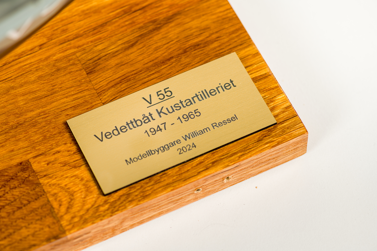 Fartygmodell av vedettbåten V 55 av plywood, plast och metall. Kamoflagemålad i grått. Notera särskilt sjökortet över Göteborgs skärgård, det var där fartyget hade sin hemvist. Däck tillverkat av 2 mm plywood. Däckshus, ankarspel, ankare, skeppsbåt, sjunkbomber och livbojar är av plast och utskrivna med 3D-skrivare. Målad med acrylfärg som antingen sprutmålats eller penselmålats.

Modellen står i plexiglasmonter på fernissad träplatta med informationsskylt.
