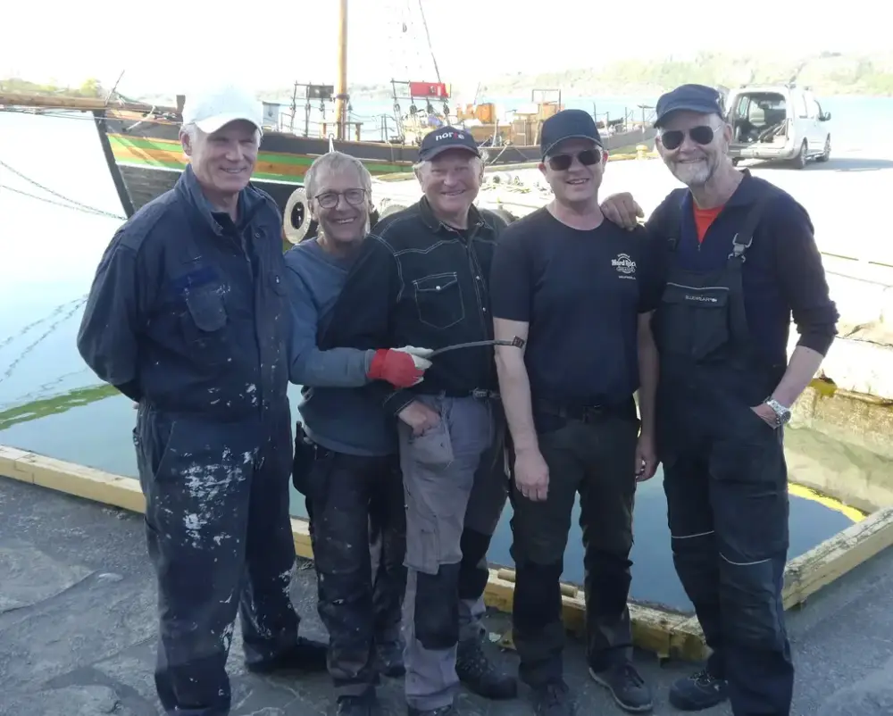 Fem menn i arbeidsklær står på kai. Seiskute i bakgrunn.