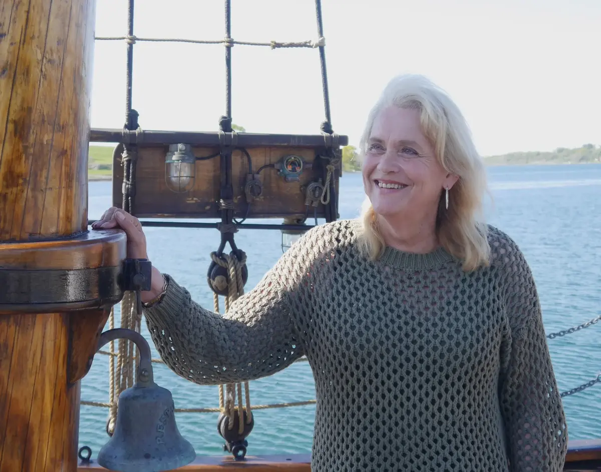 Portrett av en kvinne med lyst hår som står ved en mast med tauverk. Vann og kystlinje i bakgrunn.