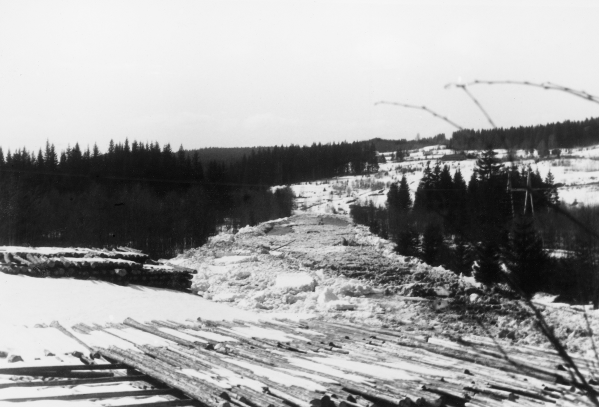 Fra Stokkeelva i daværende Snertingdal herred (seinere Gjøvik storkommune) våren 1962. Fotografiet er tatt i en periode da det var isgang i elveløpet, som i elvesvingen til høyre var tilstoppet. På elvebrinken i forgrunnen lå det tømmer i strøvelter, klart for utislag når Stokkeelva ble isfri og flomstor.