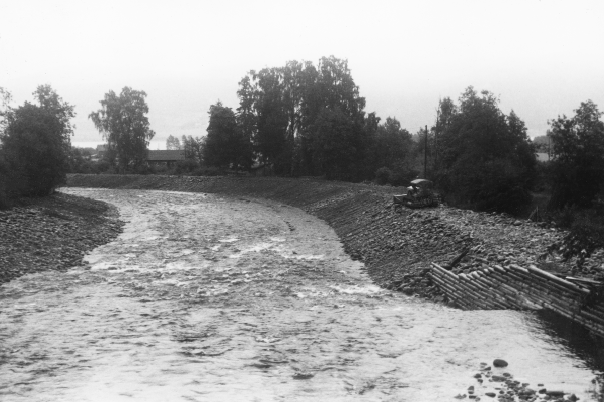 Fra den nedre delen av elva Vismunda, i nærheten av kommunesenteret i daværende Biri herred. Fotografiet ble tatt sommeren 1962, like etter at Glomma fellesfløtingsforening hadde leid bulldoser som skyflet mye av steinen fra elveløpet og opp på breddene, der den ble liggende som i voller. Bulldoseren var fortsatt i aktivitet da dette fotografiet ble tatt.  Hensikten med «elveforbedrings-» eller «bulldoserarbeidet» i var å skape bedre passasje for fløtingstømmeret. Dette bildet er tatt i medstrøms retning på et sted der det var en skådam, en skjerm som skulle hindre at fløtingstømmeret i flomperioder drev ut av elveløpet. Her var skådammen bygd som ei steinfylt tømmerkiste.