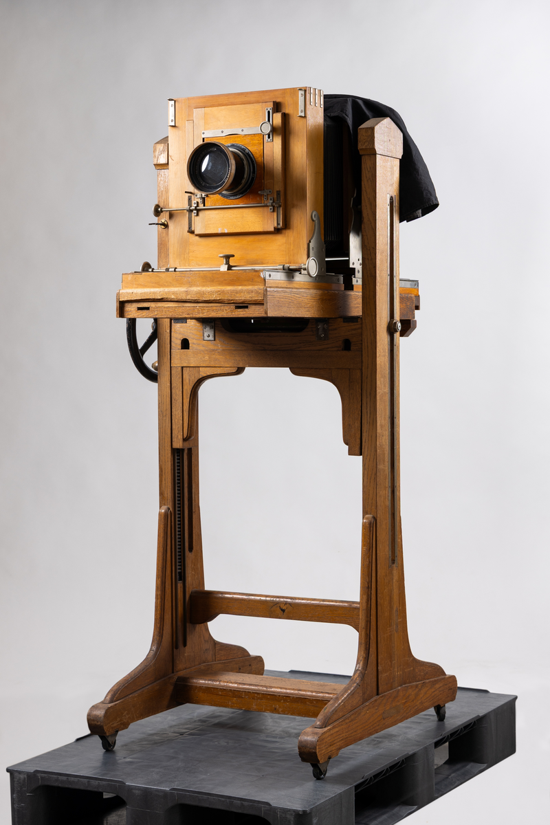Ateljékamera för glasplåtar med optik av märket Voigtländer & Sohn, Braunschweig. Linsen märkt: "Heliar 36 cm. 1:4,5. D. R. P. 124934. No 82766." Stativ av trä, höj- och sänkbart, på fyra hjul. Bälg av svart klot. Vid förvärvstillfället medföljde två svarta tygstycken, varav ett var fastsatt i kameran med häftstift. Löst linsakydd av papp.