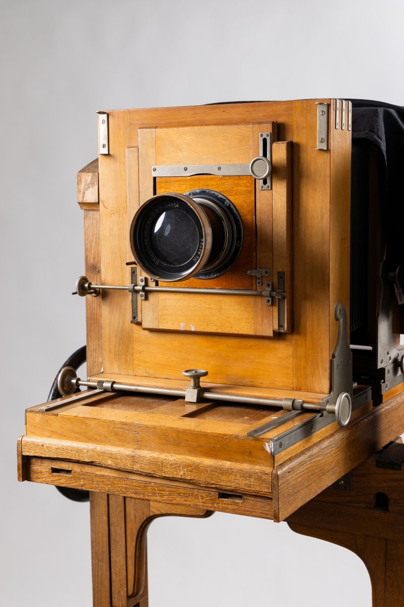 Ateljékamera för glasplåtar med optik av märket Voigtländer & Sohn, Braunschweig. Linsen märkt: "Heliar 36 cm. 1:4,5. D. R. P. 124934. No 82766." Stativ av trä, höj- och sänkbart, på fyra hjul. Bälg av svart klot. Vid förvärvstillfället medföljde två svarta tygstycken, varav ett var fastsatt i kameran med häftstift. Löst linsakydd av papp.