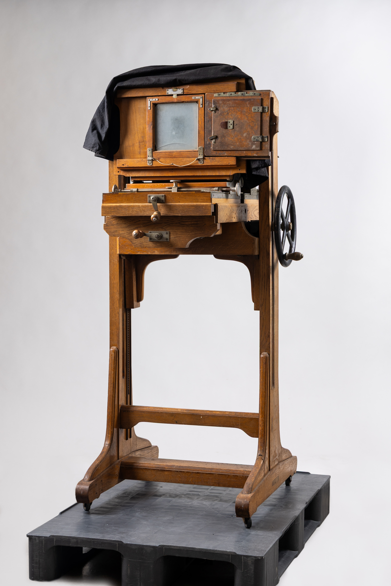 Ateljékamera för glasplåtar med optik av märket Voigtländer & Sohn, Braunschweig. Linsen märkt: "Heliar 36 cm. 1:4,5. D. R. P. 124934. No 82766." Stativ av trä, höj- och sänkbart, på fyra hjul. Bälg av svart klot. Vid förvärvstillfället medföljde två svarta tygstycken, varav ett var fastsatt i kameran med häftstift. Löst linsakydd av papp.