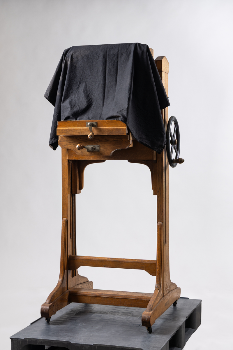 Ateljékamera för glasplåtar med optik av märket Voigtländer & Sohn, Braunschweig. Linsen märkt: "Heliar 36 cm. 1:4,5. D. R. P. 124934. No 82766." Stativ av trä, höj- och sänkbart, på fyra hjul. Bälg av svart klot. Vid förvärvstillfället medföljde två svarta tygstycken, varav ett var fastsatt i kameran med häftstift. Löst linsakydd av papp.