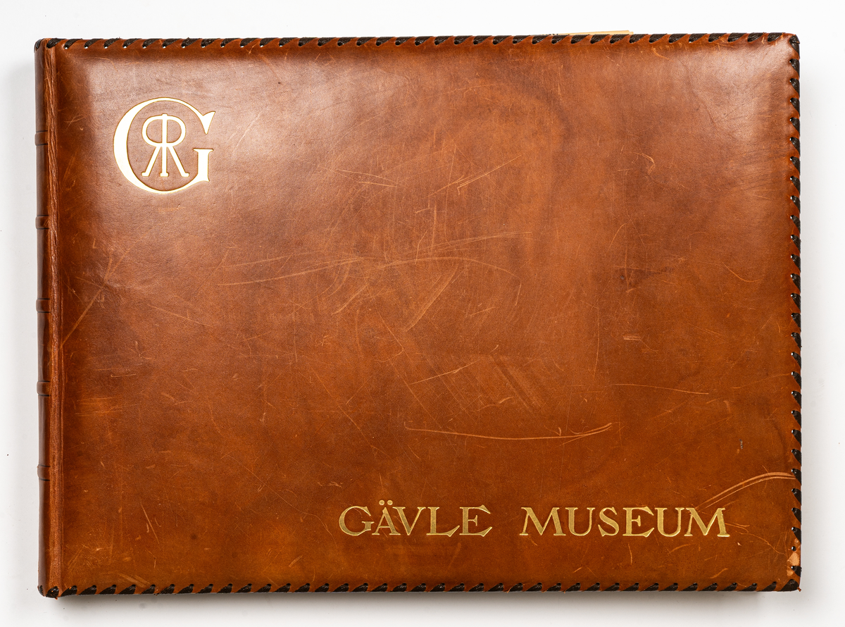 Fotoalbum med ljusbruna skinnpärmar, sydda med skinnband runt om. Text i guld: GÄVLE MUSEUM och familjen Rettigs namnchiffer inom ett G.