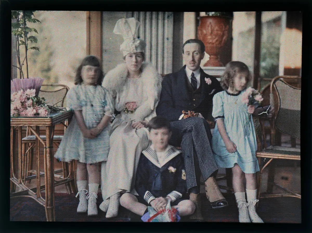 En familie i klær fra 1910-tallet er sittende i en stue, ser mot kamera. En kvinne, en mann, to jenter og en gutt. Bildet er i duse farger. 