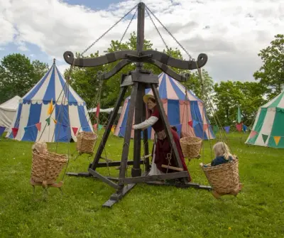 Historisk karusell i tre med flettede kurver snurret av dame med rød middelalderkjole. Barn i kurvene