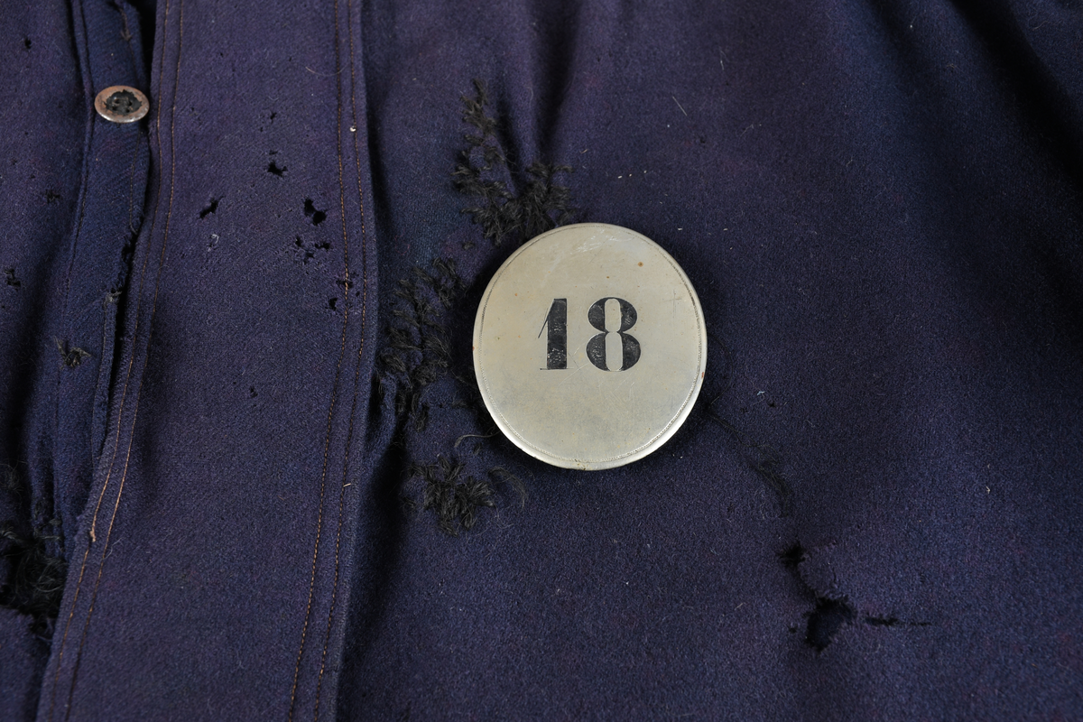 Busserull av ull, blå utførelse. Kort oppreist krage, åpning i front med tre knapper. Èn knapp nederst på hver arm til lukning. Rynket bakstykke over skuldrene. Skilt med påskrift "18" påsydd uniformen.