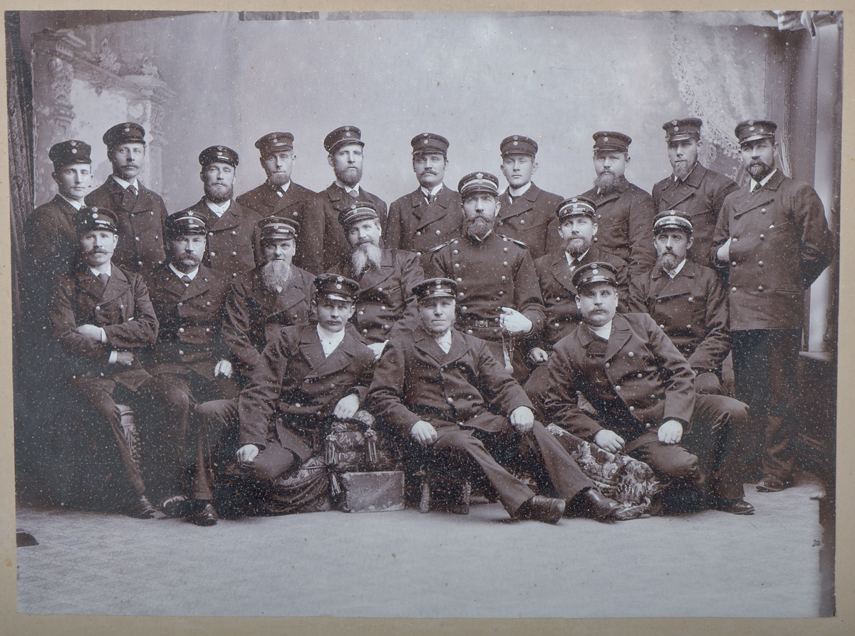 Fotografi i ram. Svartvit gruppbild i studiomiljö föreställande 20 män i uniform, identifierade som personal vid Varbergs tullkammare. Passpartout med signatur: "Mathilda Ranch Varberg".

Fotografiet är inramat i en träram, framtill förgylld och dekorerad med reliefmönster i gips. Stämpel baktill på ramen: "Göteborgs Guldlistfabrik patent". Krok och växtfibersnöre upptill för upphängning.