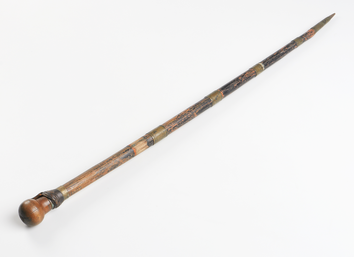 Käpp av brunmålad bambu med utdragbar värja. Rund träknopp till handtag i vilken värjan är fäst. Detaljer av mässing utmed käppen samt doppsko nedtill som avlutas i en spets. Läder virat under knoppen.