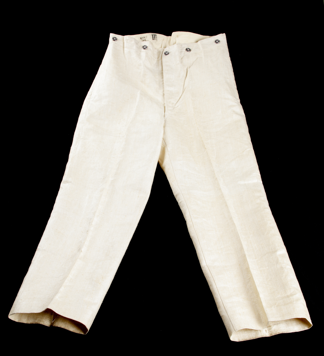 Uniformsbyxor (sommaruniform). Rak modell med pressveck, i ljusbeige, grov bomull. Knäppgylf med tre blyknappar. Två sidofickor, sju blyknappar i midjan (bärs med hängslen), sprund baktill i linningen där byxornas vidd kan regleras med ett spänne strax nedanför.