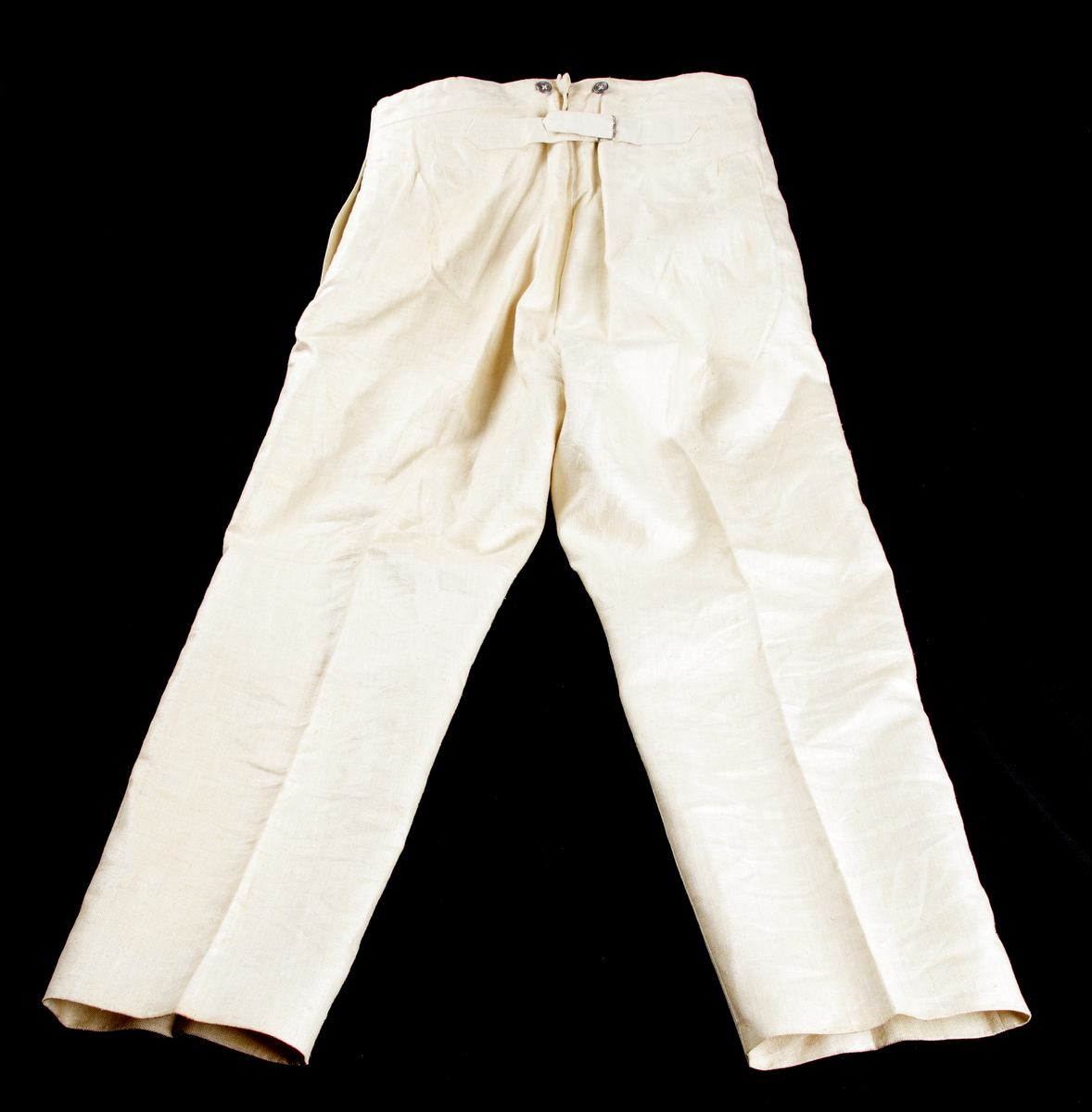 Uniformsbyxor (sommaruniform). Rak modell med pressveck, i ljusbeige, grov bomull. Knäppgylf med tre blyknappar. Två sidofickor, sju blyknappar i midjan (bärs med hängslen), sprund baktill i linningen där byxornas vidd kan regleras med ett spänne strax nedanför.